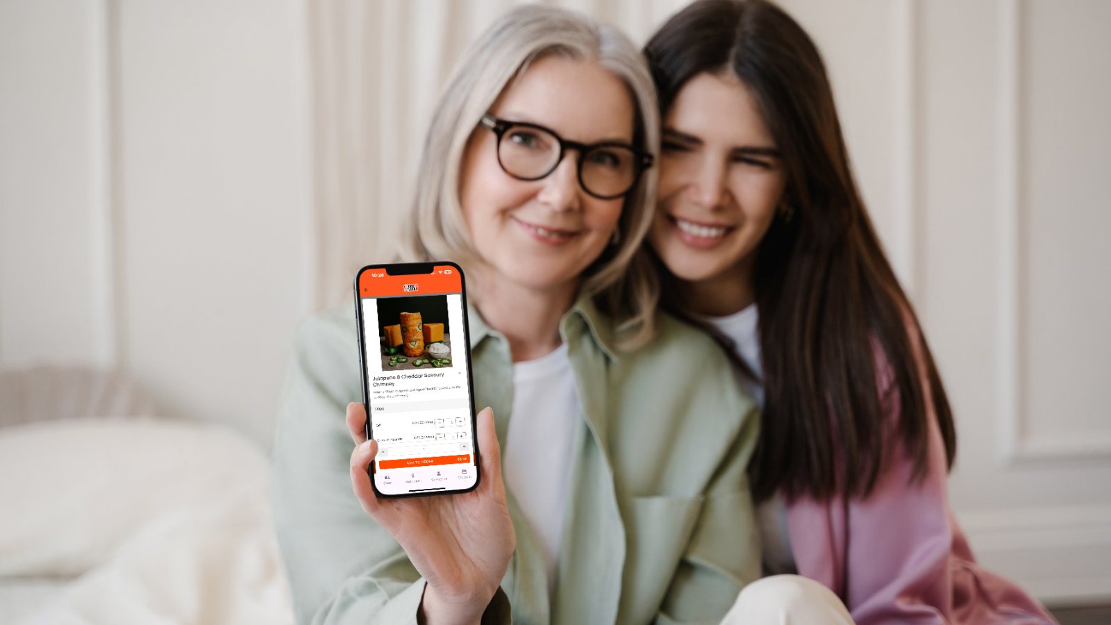 Two women, one older, one younger, sit at home smiling as they pre-order a lunch using a simple online ordering interface from Dine Local on their mobile phone.